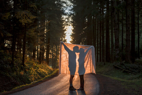 Silhouette eines Paares, das eine Decke hält und sich auf einer Landstraße im Wald küsst - WVF00907