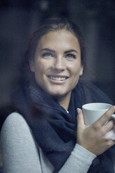 Porträt einer lachenden jungen Frau mit Kaffeetasse, die aus dem Fenster schaut - PNEF00398