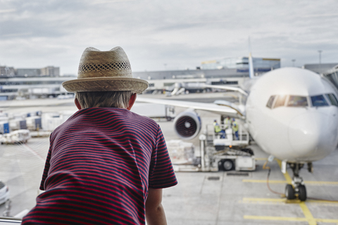 Junge mit Strohhut schaut durch ein Fenster auf ein Flugzeug auf dem Vorfeld, lizenzfreies Stockfoto