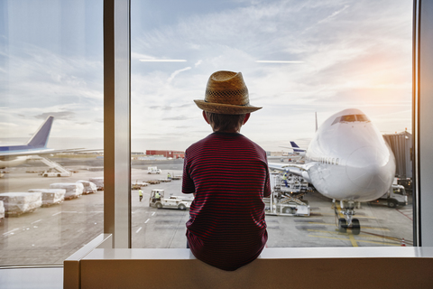 Junge mit Strohhut schaut durch ein Fenster auf ein Flugzeug auf dem Vorfeld, lizenzfreies Stockfoto