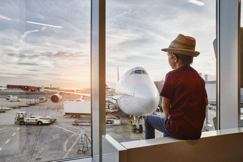 Junge mit Strohhut schaut durch ein Fenster auf ein Flugzeug auf dem Vorfeld, lizenzfreies Stockfoto