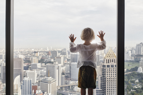 Thailand, Bangkok, kleines Mädchen schaut durch ein Fenster auf eine Stadtlandschaft, lizenzfreies Stockfoto