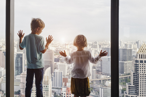 Thailand, Bangkok, Junge und kleines Mädchen schauen durch ein Fenster auf eine Stadtlandschaft, lizenzfreies Stockfoto