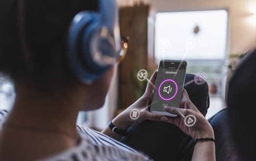 Woman sitting at home with headphones, using smartphone as remote control - UUF12530