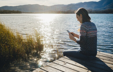 Frau sitzt auf einer Holzplattform an einem See bei Sonnenuntergang und benutzt ein Smartphone - GEMF01856
