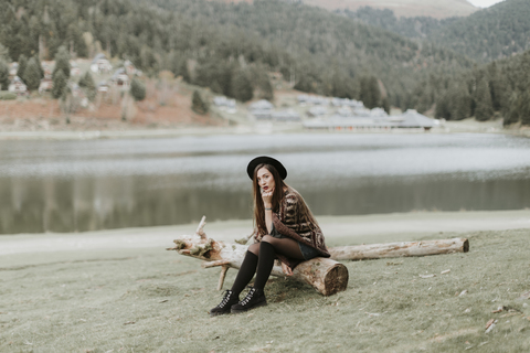 Fashionable young woman wearing hat and poncho sitting on log on a meadow stock photo