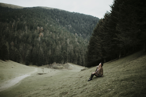 Junge Frau sitzt auf einer Wiese am Waldrand und schaut nach oben, lizenzfreies Stockfoto