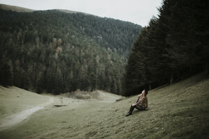 Junge Frau sitzt auf einer Wiese am Waldrand und schaut nach oben - OCAF00080