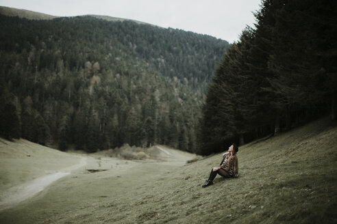 Young woman sitting on a meadow near forest edge looking up - OCAF00080