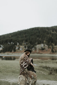 Fashionable young woman wearing hat and poncho sitting on a rock - OCAF00081