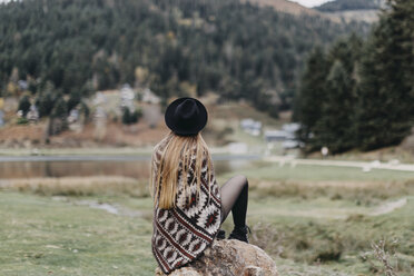 Rückenansicht einer modischen jungen Frau mit Hut und Poncho, die auf einem Felsen sitzt - OCAF00082