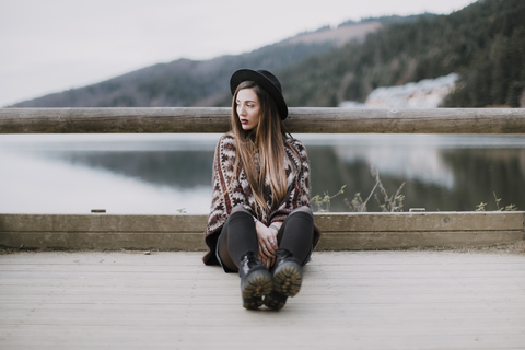 Portrait of fashionable young woman wearing hat and poncho sitting on wood bridge stock photo