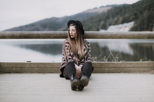 Porträt einer modischen jungen Frau mit Hut und Poncho auf einer Holzbrücke sitzend - OCAF00083