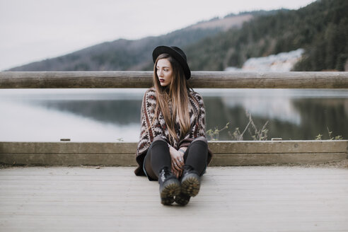 Portrait of fashionable young woman wearing hat and poncho sitting on wood bridge - OCAF00083
