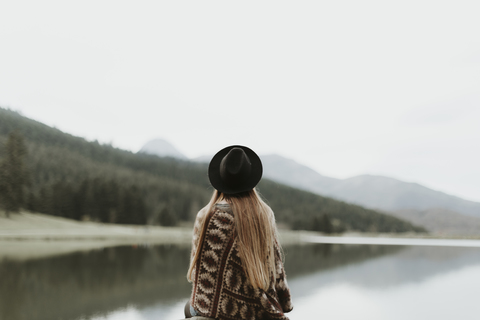 Rückenansicht einer jungen Frau mit Hut und Poncho, die vor einem See sitzt, lizenzfreies Stockfoto