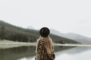 Rückenansicht einer jungen Frau mit Hut und Poncho, die vor einem See sitzt - OCAF00084