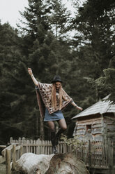 Fashionable young woman wearing hat and poncho balancing on tree trunk - OCAF00087