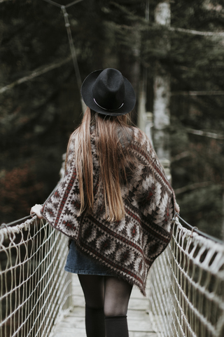 Back view of fashionable young woman wearing hat and poncho walking on suspension bridge stock photo