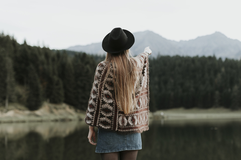 Rückenansicht einer modischen jungen Frau mit Hut und Poncho, die vor einem See steht, lizenzfreies Stockfoto