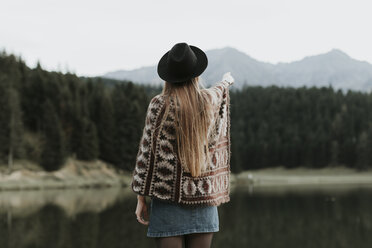 Back view of fashionable young woman wearing hat and poncho standing in front of a lake - OCAF00090