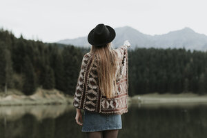 Rückenansicht einer modischen jungen Frau mit Hut und Poncho, die vor einem See steht - OCAF00090