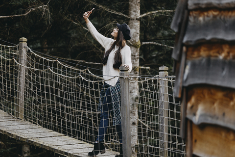 Junge Frau auf einer Hängebrücke am Baumhaus im Wald macht ein Selfie, lizenzfreies Stockfoto