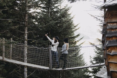 Zwei junge Frauen auf einer Hängebrücke am Baumhaus im Wald machen ein Selfie - OCAF00097