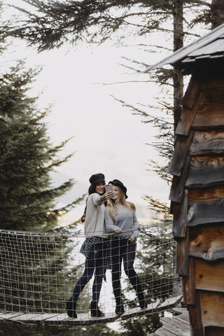 Zwei junge Frauen auf einer Hängebrücke am Baumhaus im Wald machen ein Selfie, lizenzfreies Stockfoto