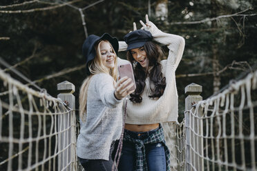 Zwei glückliche junge Frauen auf einer Hängebrücke machen ein Selfie - OCAF00099