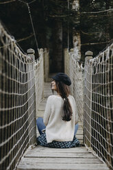 Smiling young woman sitting on a suspension bridge - OCAF00104