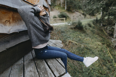 Glückliche junge Frau sitzt an einem Holzhaus in der Natur - OCAF00111