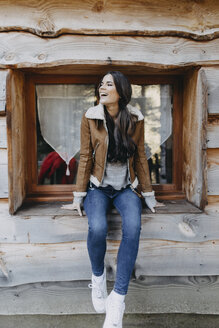 Glückliche junge Frau, die auf der Fensterbank eines Holzhauses sitzt - OCAF00113