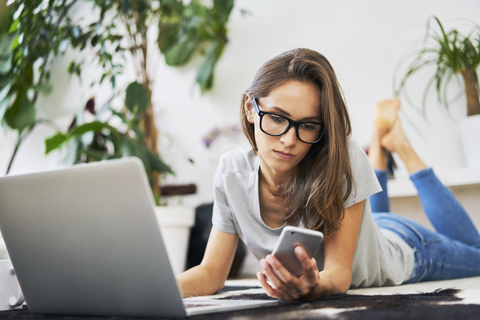 Young woman at home lying on the floor with cell phone and laptop stock photo