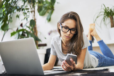 Young woman at home lying on the floor with cell phone and laptop - BSZF00147