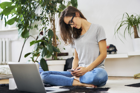 Junge Frau zu Hause, die auf dem Boden sitzt und ihr Smartphone benutzt, lizenzfreies Stockfoto