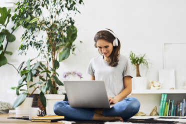 Junge Frau zu Hause, die auf dem Boden sitzt, einen Laptop benutzt und Musik hört - BSZF00150