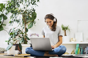 Junge Frau zu Hause, die auf dem Boden sitzt, einen Laptop benutzt und Musik hört - BSZF00150