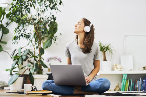 Entspannte junge Frau, die zu Hause auf dem Boden sitzt, einen Laptop benutzt und Musik hört, lizenzfreies Stockfoto