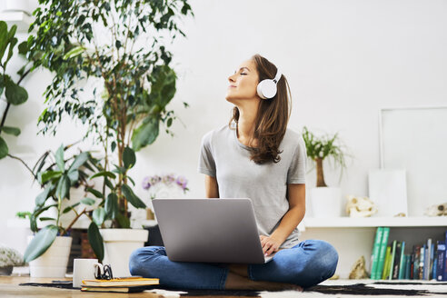 Entspannte junge Frau, die zu Hause auf dem Boden sitzt, einen Laptop benutzt und Musik hört - BSZF00151