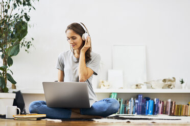 Entspannte junge Frau, die zu Hause auf dem Boden sitzt, einen Laptop benutzt und Musik hört - BSZF00152