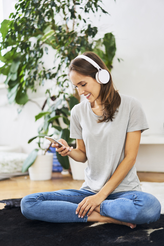 Smiling young woman sitting on the floor with headphones and cell phone stock photo