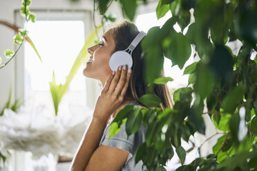 Lächelnde junge Frau mit Kopfhörern, die in einer Zimmerpflanze Musik hört - BSZF00157