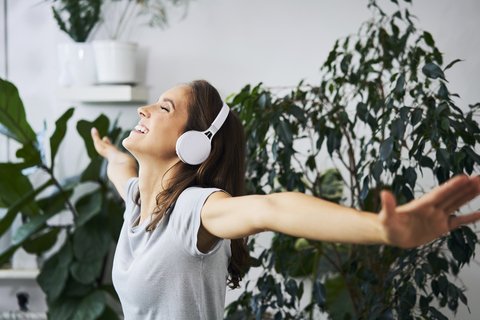 Glückliche junge Frau mit ausgestreckten Armen, die in einer Zimmerpflanze Musik hört, lizenzfreies Stockfoto