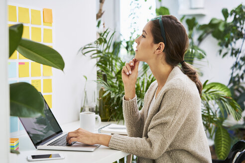 Junge Frau mit Laptop auf dem Schreibtisch und Blick auf Haftnotizen an der Wand - BSZF00164