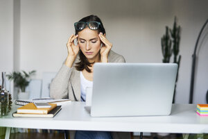 Junge Frau zu Hause mit Laptop auf dem Schreibtisch, der ihre Schläfen berührt - BSZF00168