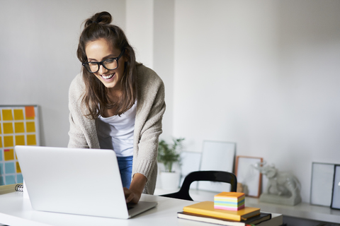 Happy young woman at home using laptop on desk stock photo