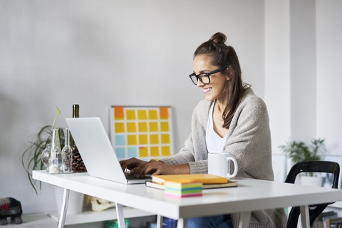 Lächelnde junge Frau zu Hause mit Laptop auf dem Schreibtisch, lizenzfreies Stockfoto