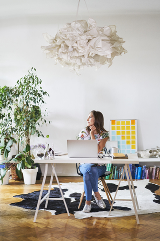 Junge Frau zu Hause mit Laptop auf dem Schreibtisch mit Blick zur Seite, lizenzfreies Stockfoto