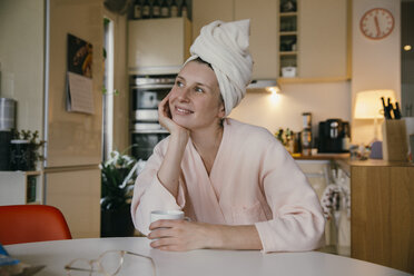 Porträt einer lächelnden Frau mit Handtuch-Turban, die mit einer Tasse Kaffee am Küchentisch sitzt - MFF04374