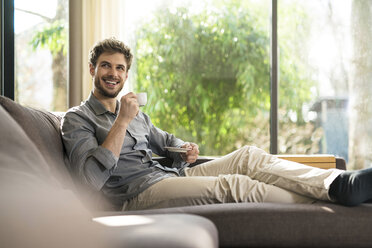 Laughing man relaxing on couch at home drinking coffee - SBOF01259